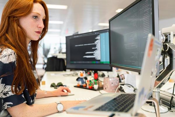 Woman working at an office desk reviewing computer code on two monitors