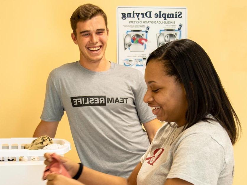 Two students smile while folding laundry in a dorm laundry room.